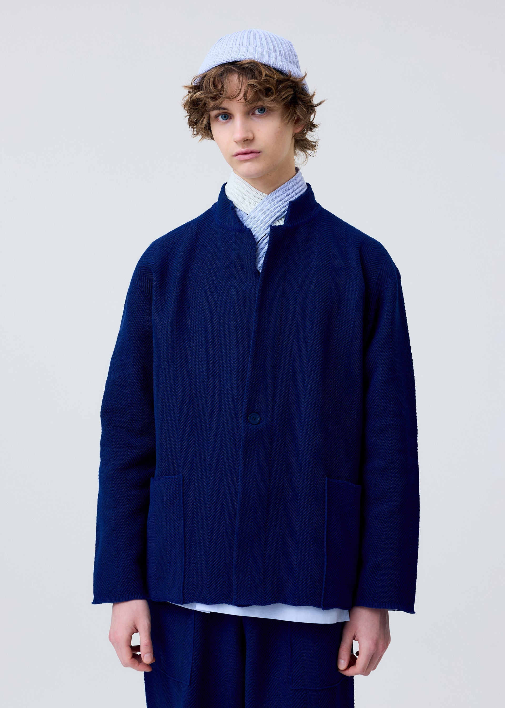 Zoomed view A person with curly hair wears the HOMME PLISSÉ ISSEY MIYAKE INLAID KNIT navy blue jacket and matching pants, a light blue shirt, and a light blue beanie, standing against a plain light background.