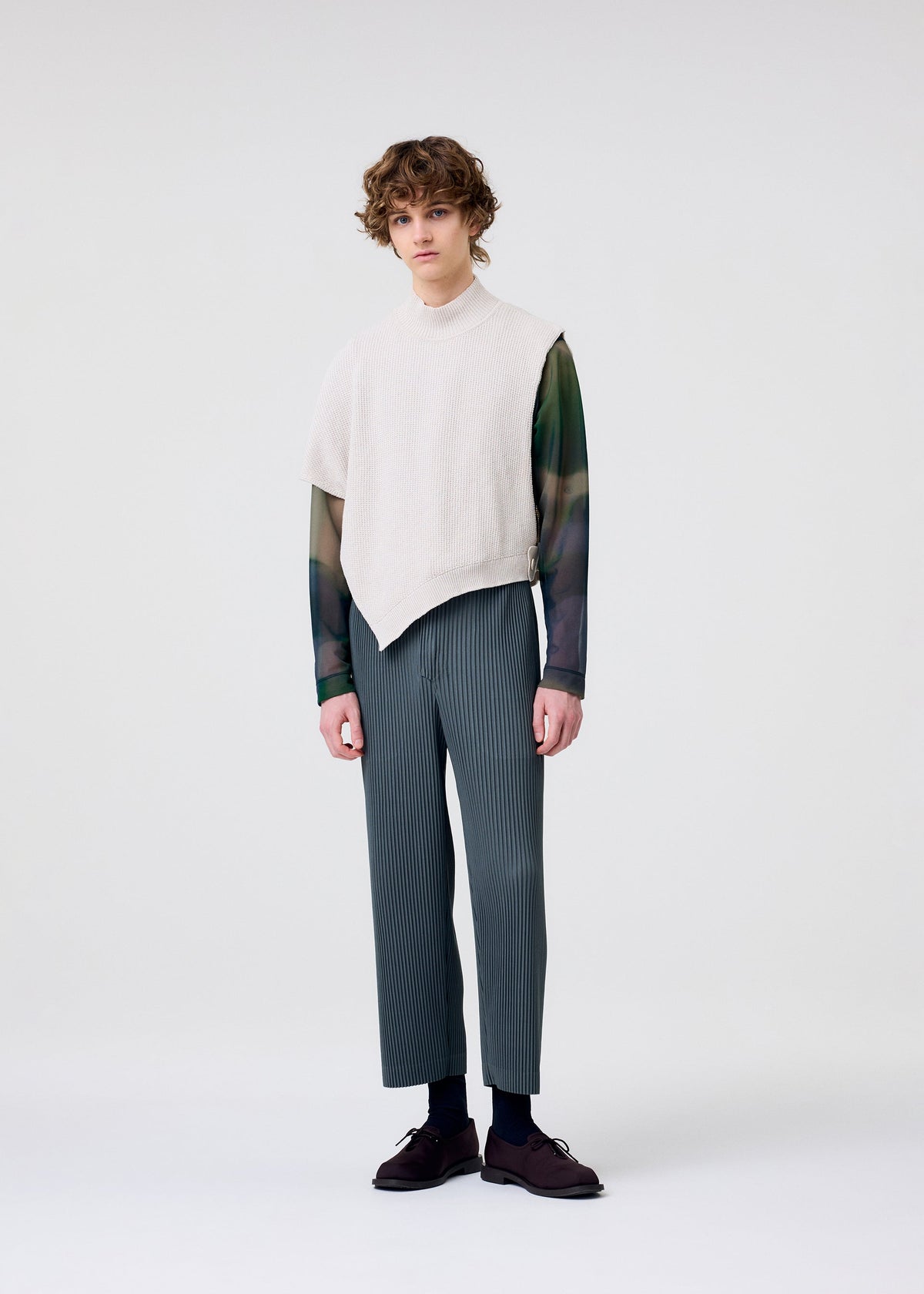 A person stands against a plain white background, wearing the HOMME PLISSÉ ISSEY MIYAKE WAFER KNIT over a sheer long-sleeve shirt and gray pinstriped pants with dark shoes, showcasing a modern layered look while gazing neutrally at the camera. view 6 of 8