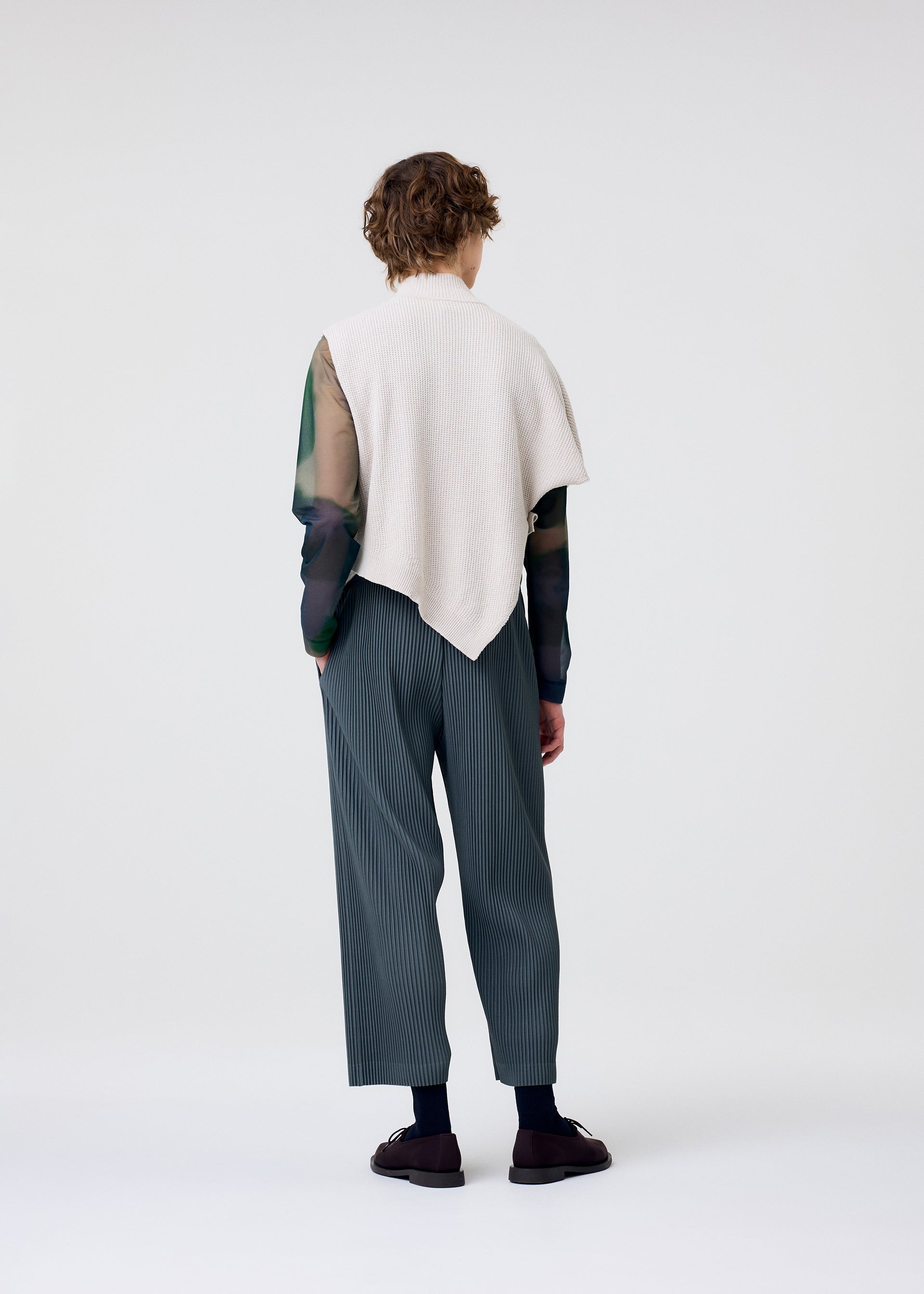 Zoomed view A person with short, curly hair showcases the HOMME PLISSÉ ISSEY MIYAKE WAFER KNIT top in cream over a sheer green long-sleeve, paired with loose striped gray pants and black shoes against a simple, light background.