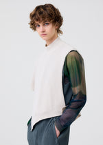 Wearing the HOMME PLISSÉ ISSEY MIYAKE WAFER KNIT top over a sheer green and black shirt with pleated gray pants, a person with curly brown hair showcases a chic layered look against a plain light background.