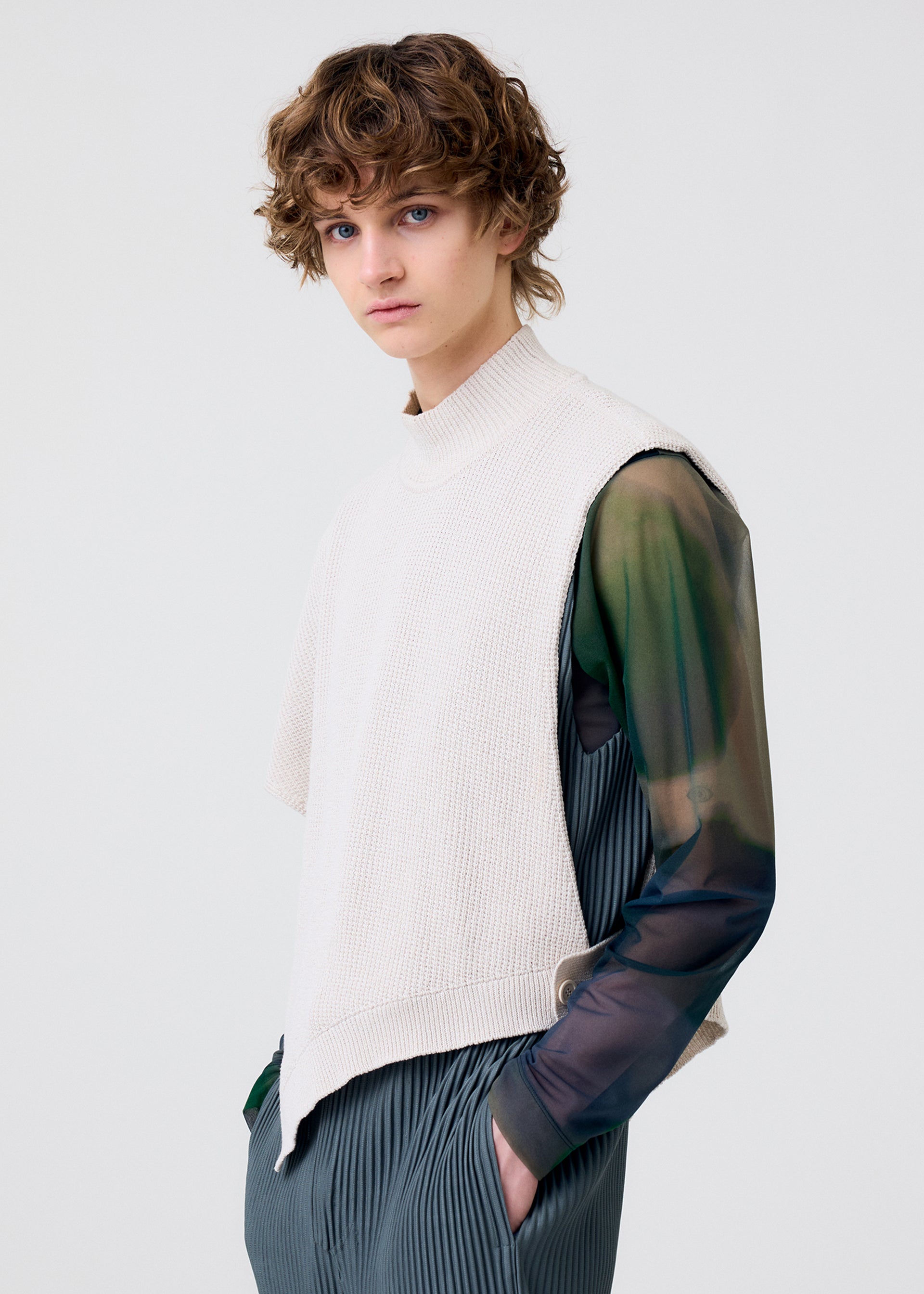 Zoomed view Wearing the HOMME PLISSÉ ISSEY MIYAKE WAFER KNIT top over a sheer green and black shirt with pleated gray pants, a person with curly brown hair showcases a chic layered look against a plain light background.