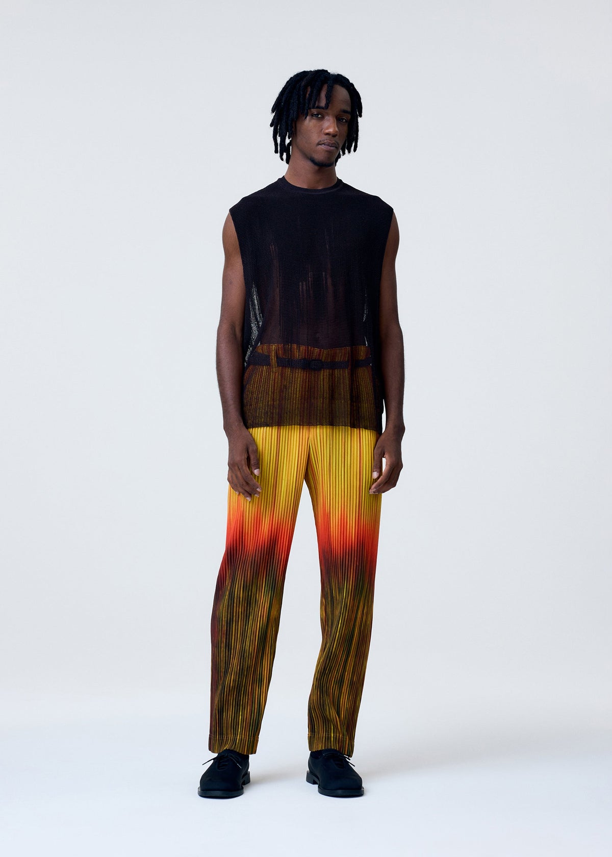 A man wears the BRUSH CLOSE-UP KNIT by HOMME PLISSÉ ISSEY MIYAKE, a sleeveless black sheer vest, paired with pleated yellow-orange ombré gradient pants and black shoes against a plain background. view 5 of 7