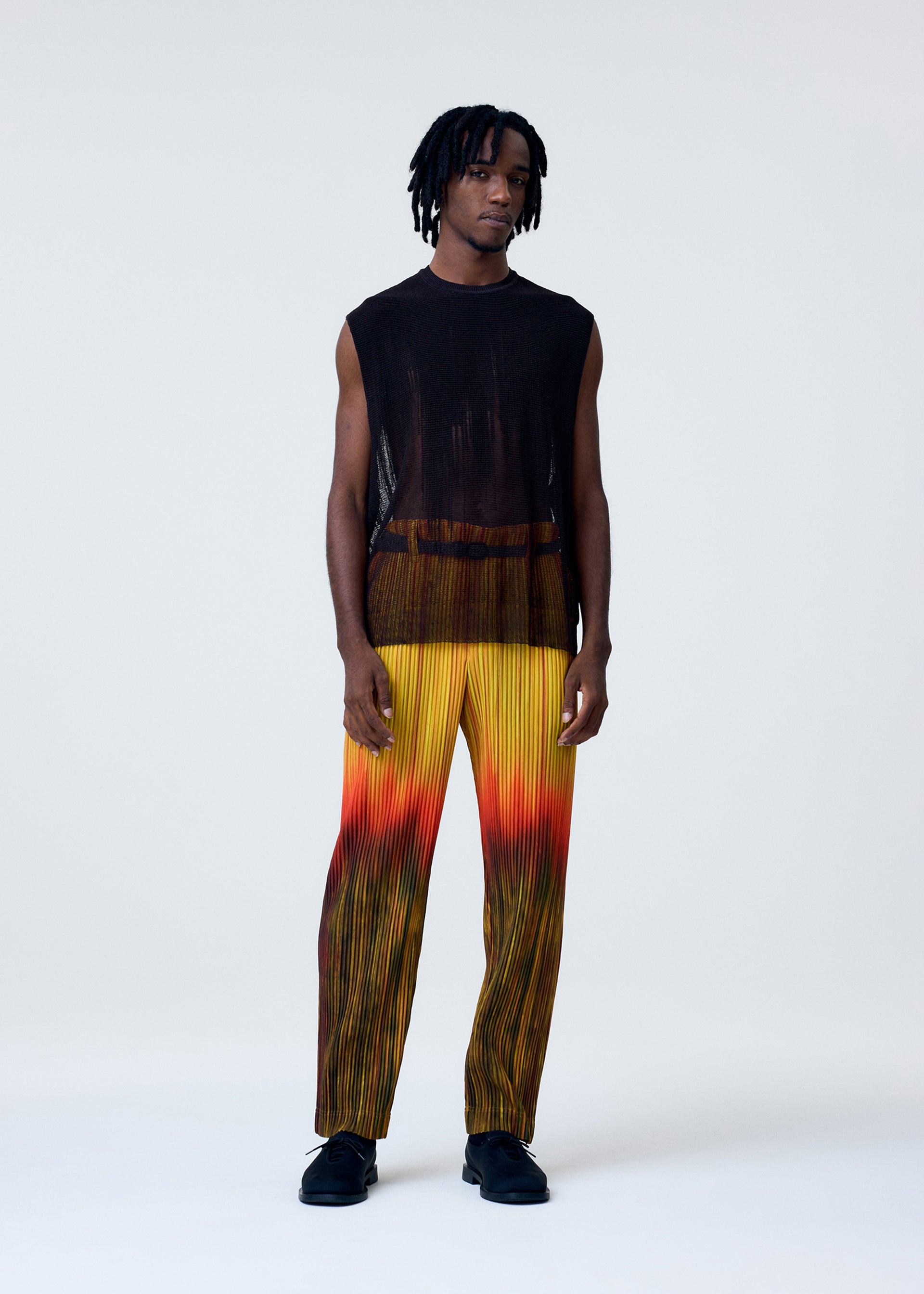 Zoomed view A man wears the BRUSH CLOSE-UP KNIT by HOMME PLISSÉ ISSEY MIYAKE, a sleeveless black sheer vest, paired with pleated yellow-orange ombré gradient pants and black shoes against a plain background.