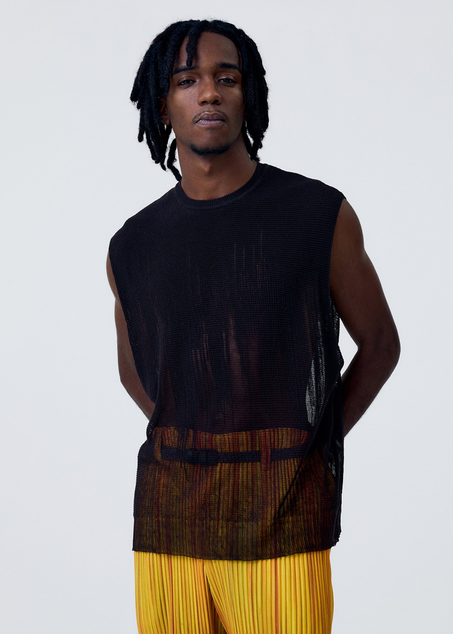 Zoomed view Wearing the HOMME PLISSÉ ISSEY MIYAKE BRUSH CLOSE-UP KNIT, a person with short dreadlocks stands against a plain light background, layering the round-neck sheer black vest over yellow pleated pants, hands behind their back.