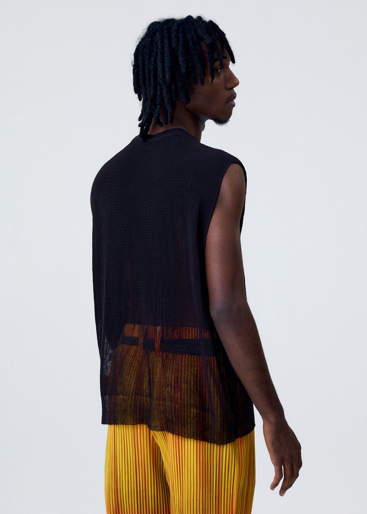 A person with short, twisted hair wears the HOMME PLISSÉ ISSEY MIYAKE BRUSH CLOSE-UP KNIT vest layered under a sleeveless, sheer black top and yellow pleated pants, standing turned slightly away against a plain light background. view 7 of 7