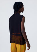 A person with short, twisted hair wears the HOMME PLISSÉ ISSEY MIYAKE BRUSH CLOSE-UP KNIT vest layered under a sleeveless, sheer black top and yellow pleated pants, standing turned slightly away against a plain light background.