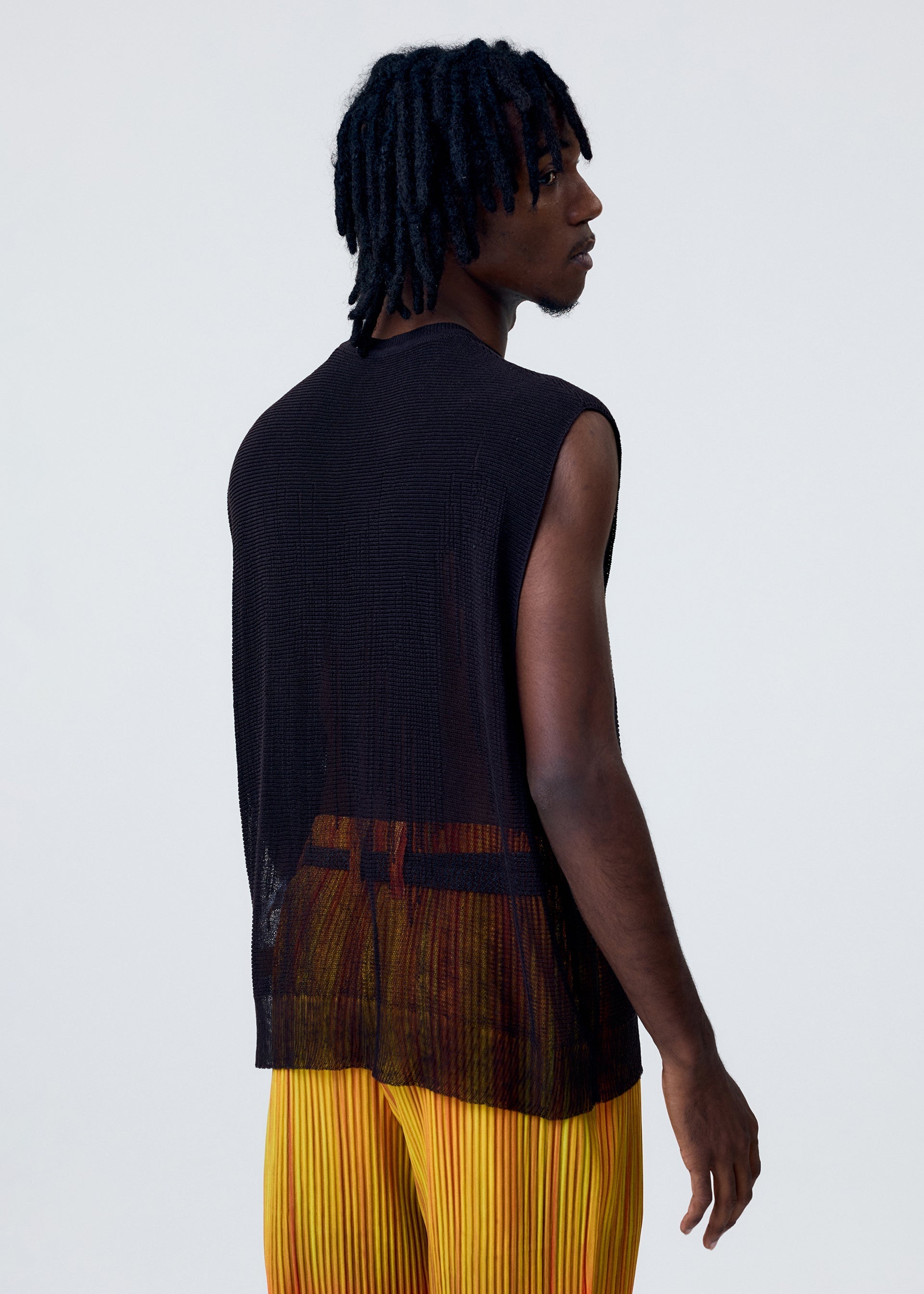 Zoomed view A person with short, twisted hair wears the HOMME PLISSÉ ISSEY MIYAKE BRUSH CLOSE-UP KNIT vest layered under a sleeveless, sheer black top and yellow pleated pants, standing turned slightly away against a plain light background.