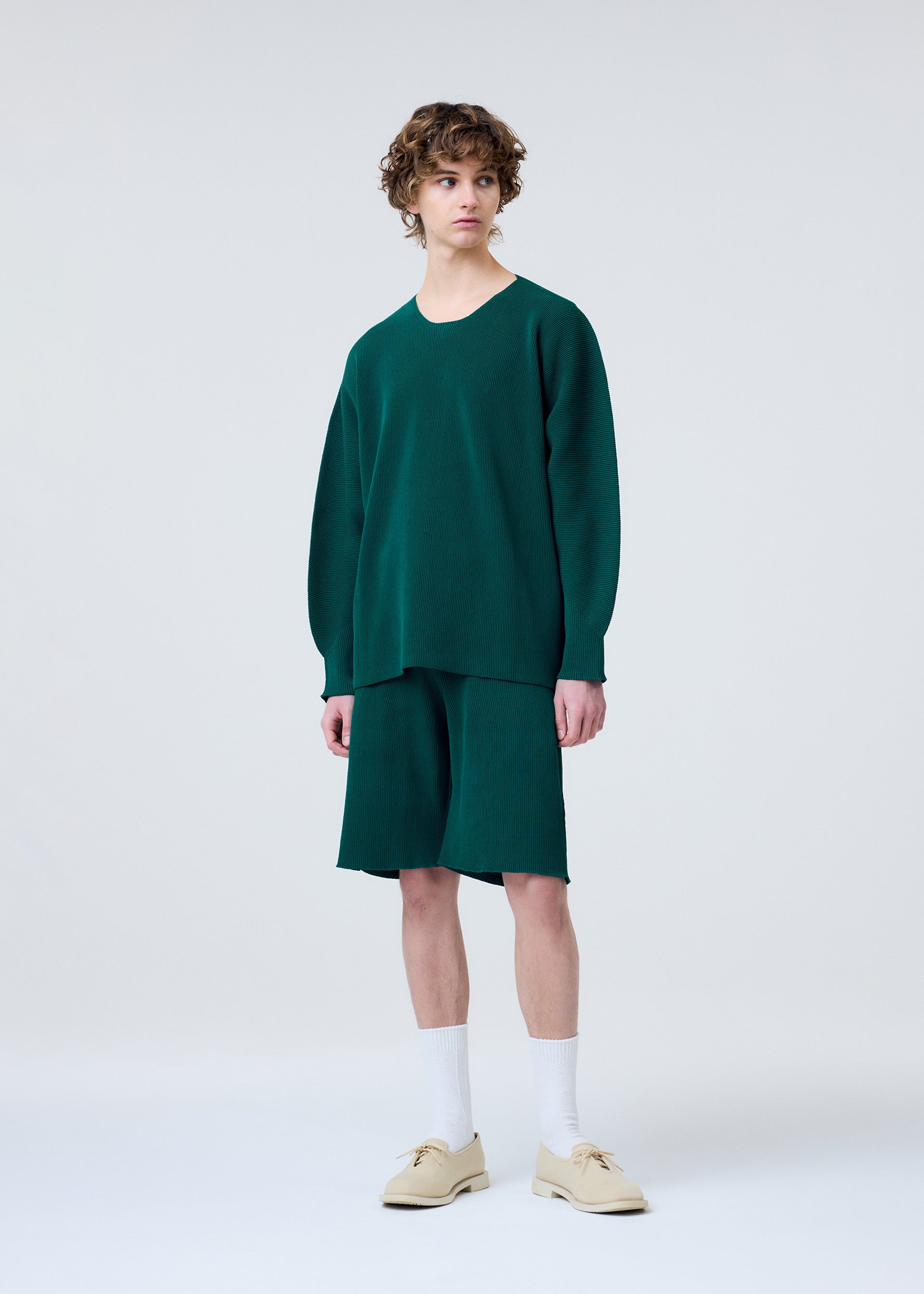 Zoomed view A person wearing a loose-fitting green long-sleeve shirt and matching HOMME PLISSÉ ISSEY MIYAKE BASIC KNIT pleated jersey shorts, paired with white socks and beige shoes, stands against a plain light gray background.