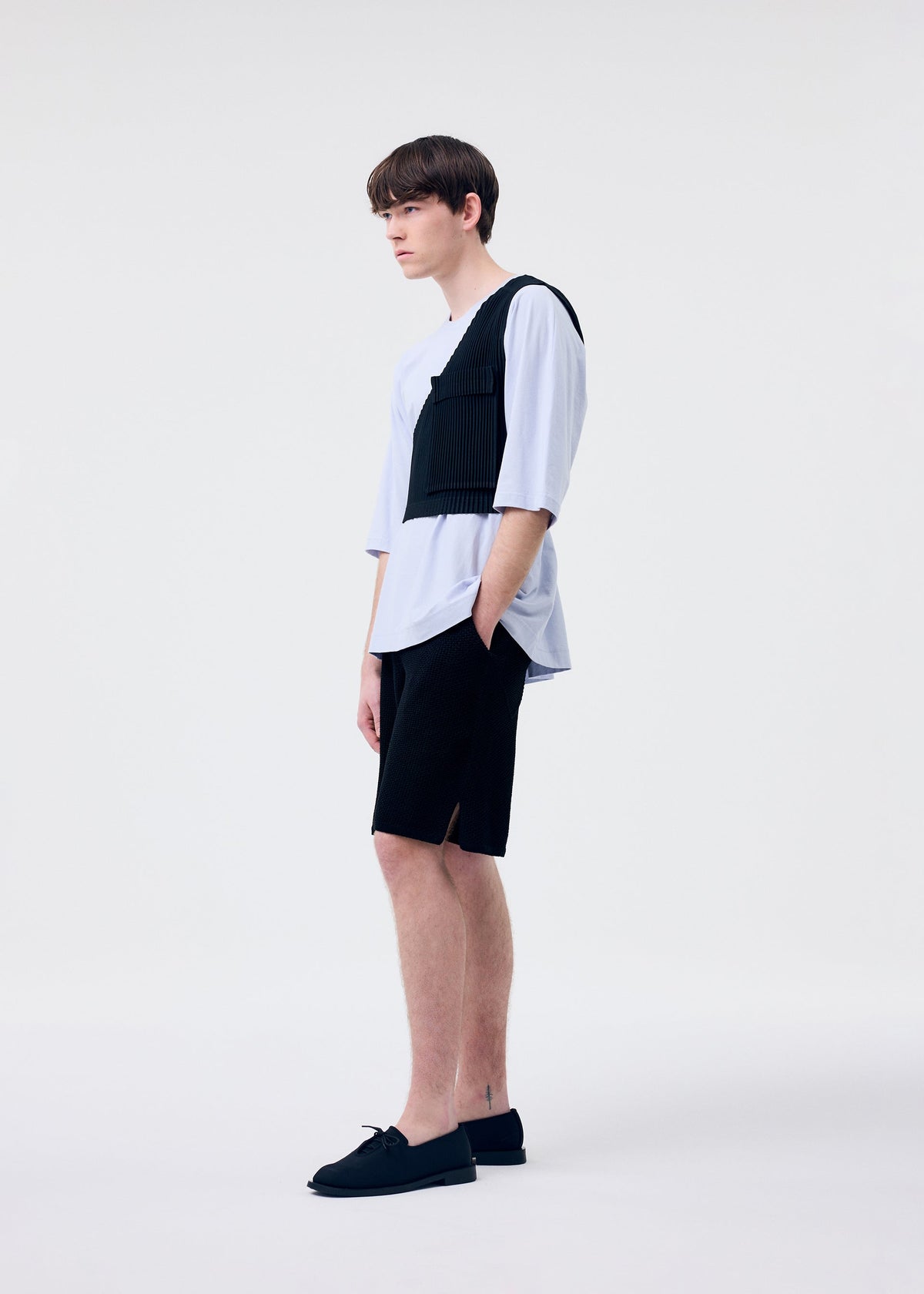 A young man stands in profile against a white background, wearing HOMME PLISSÉ ISSEY MIYAKE's RUSTIC KNIT vest over a light blue shirt, paired with black knit shorts and slip-on shoes. His hands rest in his pockets, and he has a neutral expression. view 8 of 8