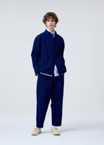 A person with wavy brown hair wears the HOMME PLISSÉ ISSEY MIYAKE INLAID KNIT dark blue suit, paired with a white shirt and beige shoes, standing against a plain light background.