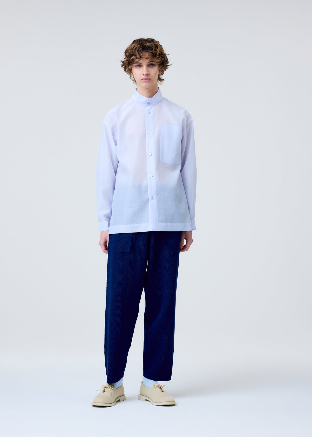 A person with short, curly hair stands against a plain background, wearing the HOMME PLISSÉ ISSEY MIYAKE INLAID KNIT navy blue pants with a subtle herringbone pattern, a loose white shirt with a large chest pocket, and beige shoes. view 7 of 7