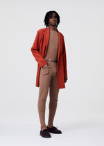 Wearing the KNIT UNRAVELED set by HOMME PLISSÉ ISSEY MIYAKE, a person stands against a plain background in a rust coat over a beige fitted top and pants, styled with sheer tights, a belt, and dark brown shoes.