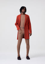 A person with medium-length dreadlocks wears the KNIT UNRAVELED coat by HOMME PLISSÉ ISSEY MIYAKE over a tan outfit with sheer tights, a matching belt, and shoes, standing against a plain white background.