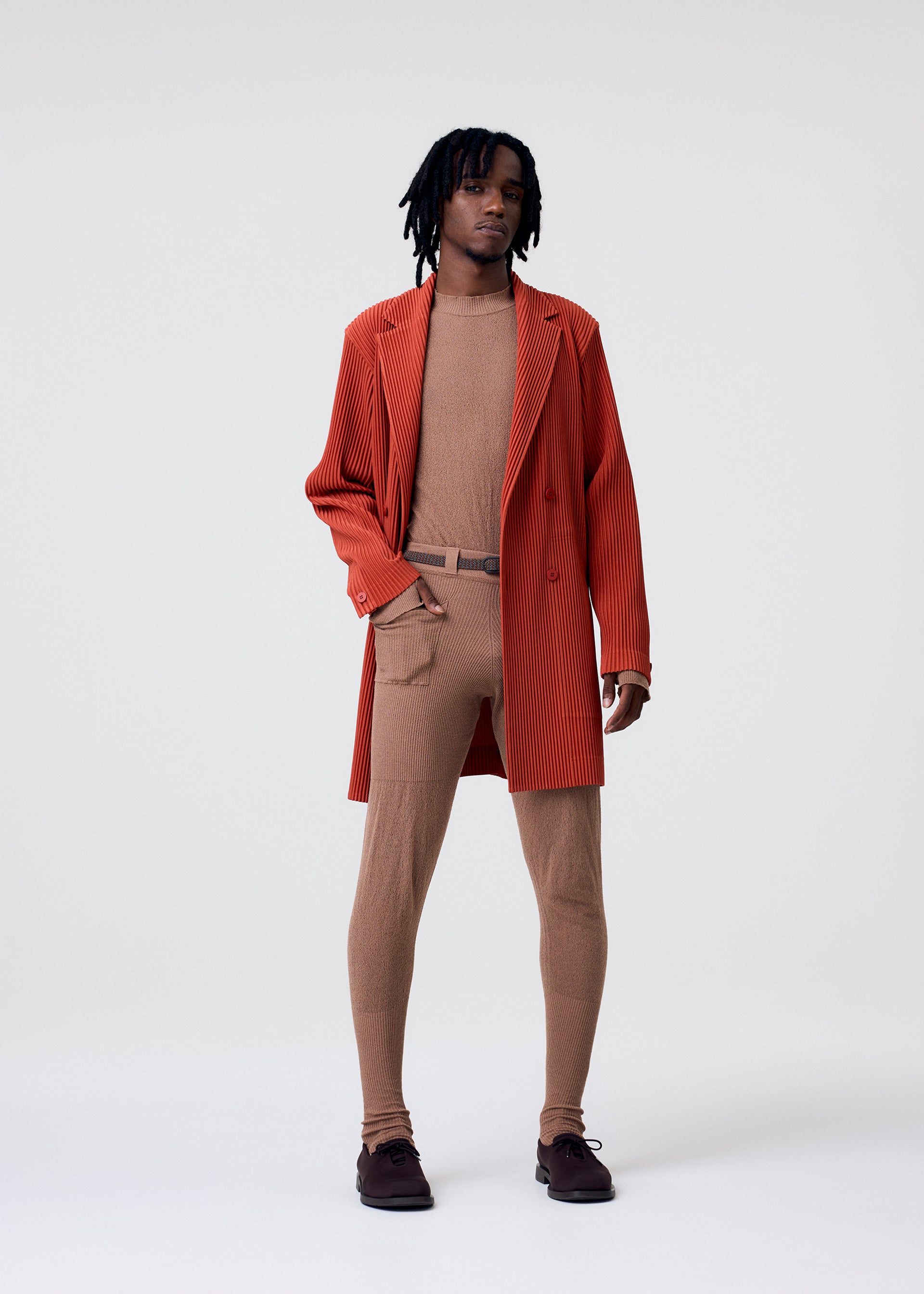 Zoomed view A person with medium-length dreadlocks wears the KNIT UNRAVELED coat by HOMME PLISSÉ ISSEY MIYAKE over a tan outfit with sheer tights, a matching belt, and shoes, standing against a plain white background.