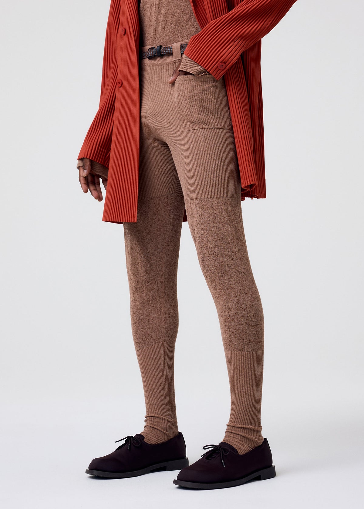 A person models HOMME PLISSÉ ISSEY MIYAKE's KNIT UNRAVELED ribbed beige leggings, layered over sheer tights with a matching top, slim belt, long red pleated coat, and dark brown lace-ups against a plain light background. Face not shown. view 8 of 8