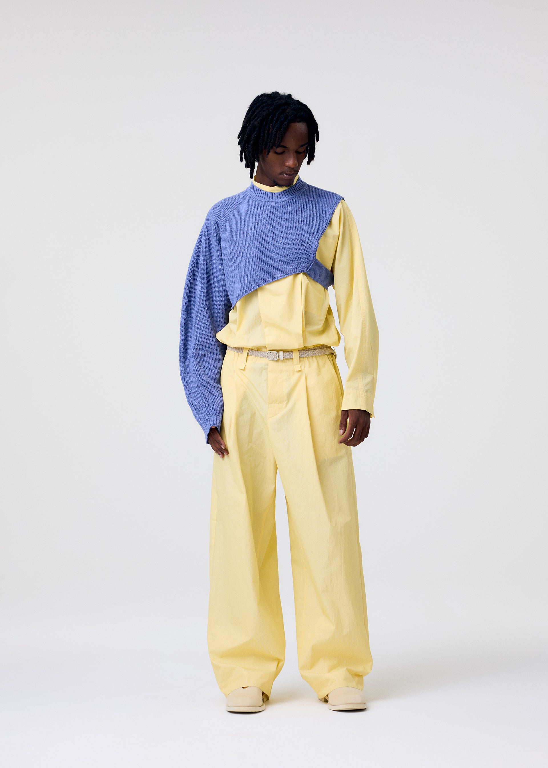Zoomed view A person stands against a plain background wearing HOMME PLISSÉ ISSEY MIYAKE's WAFER KNIT top draped over one shoulder, paired with loose-fitting yellow pants and shirt, looking down with hands by their sides, showcasing a modern layered style.