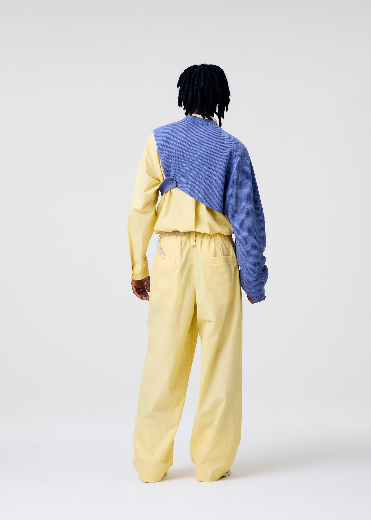 A person faces away in the HOMME PLISSÉ ISSEY MIYAKE WAFER KNIT: a light yellow set with loose pants, matching shirt, and an asymmetrical knit draped over one shoulder. The plain white background highlights the minimal layered style. view 7 of 8