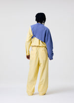 A person faces away in the HOMME PLISSÉ ISSEY MIYAKE WAFER KNIT: a light yellow set with loose pants, matching shirt, and an asymmetrical knit draped over one shoulder. The plain white background highlights the minimal layered style.