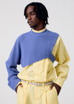A person with medium-length locs wears HOMME PLISSÉ ISSEY MIYAKE's WAFER KNIT, a blue asymmetrical top layered over a yellow button-up shirt and pants, posing hands-in-pockets against a plain light background.