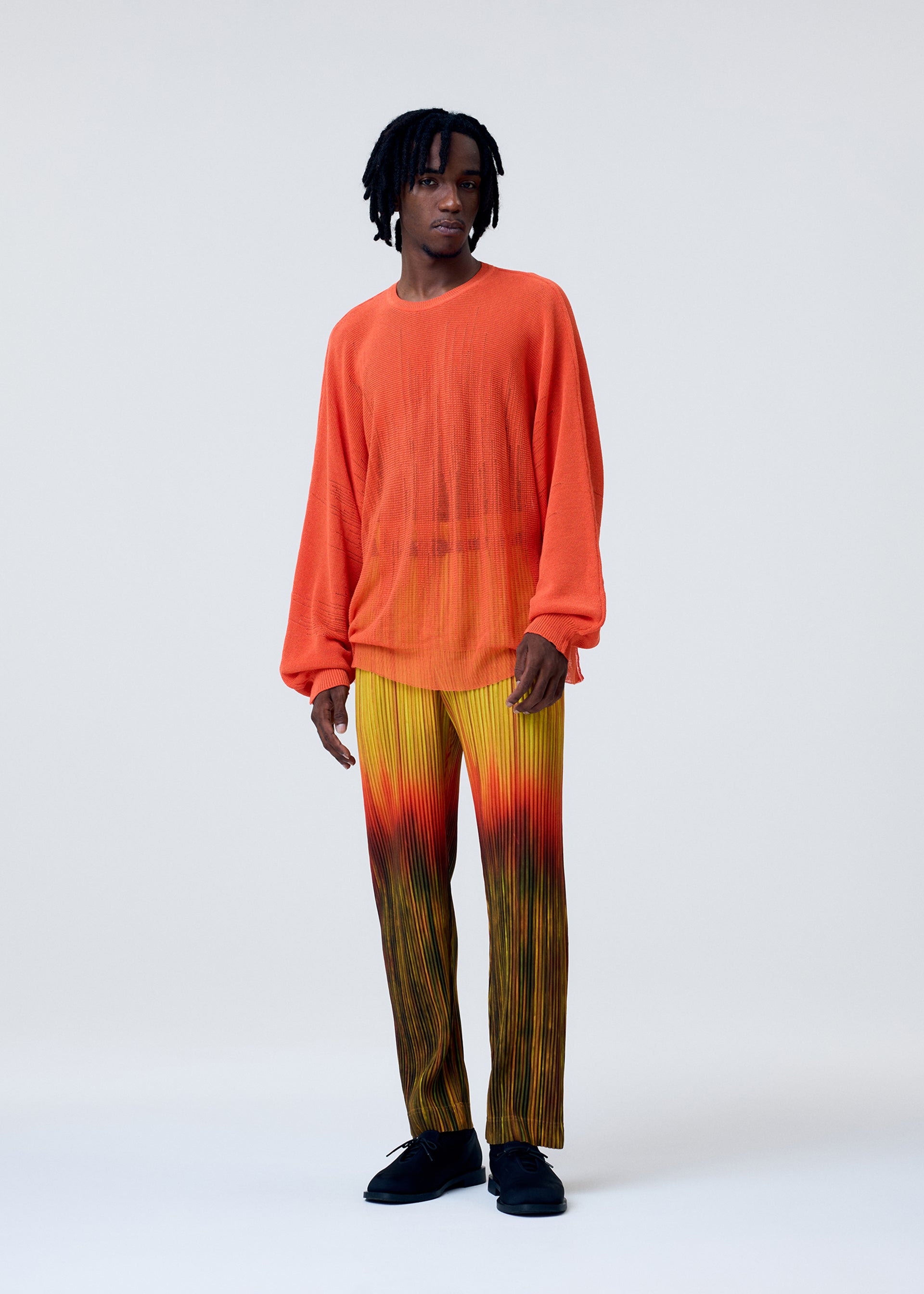 Zoomed view A person stands against a plain background wearing the BRUSH CLOSE-UP KNIT by HOMME PLISSÉ ISSEY MIYAKE—an orange, sheer, extra-long-staple cotton long-sleeved top—and pleated yellow gradient pants with black shoes.