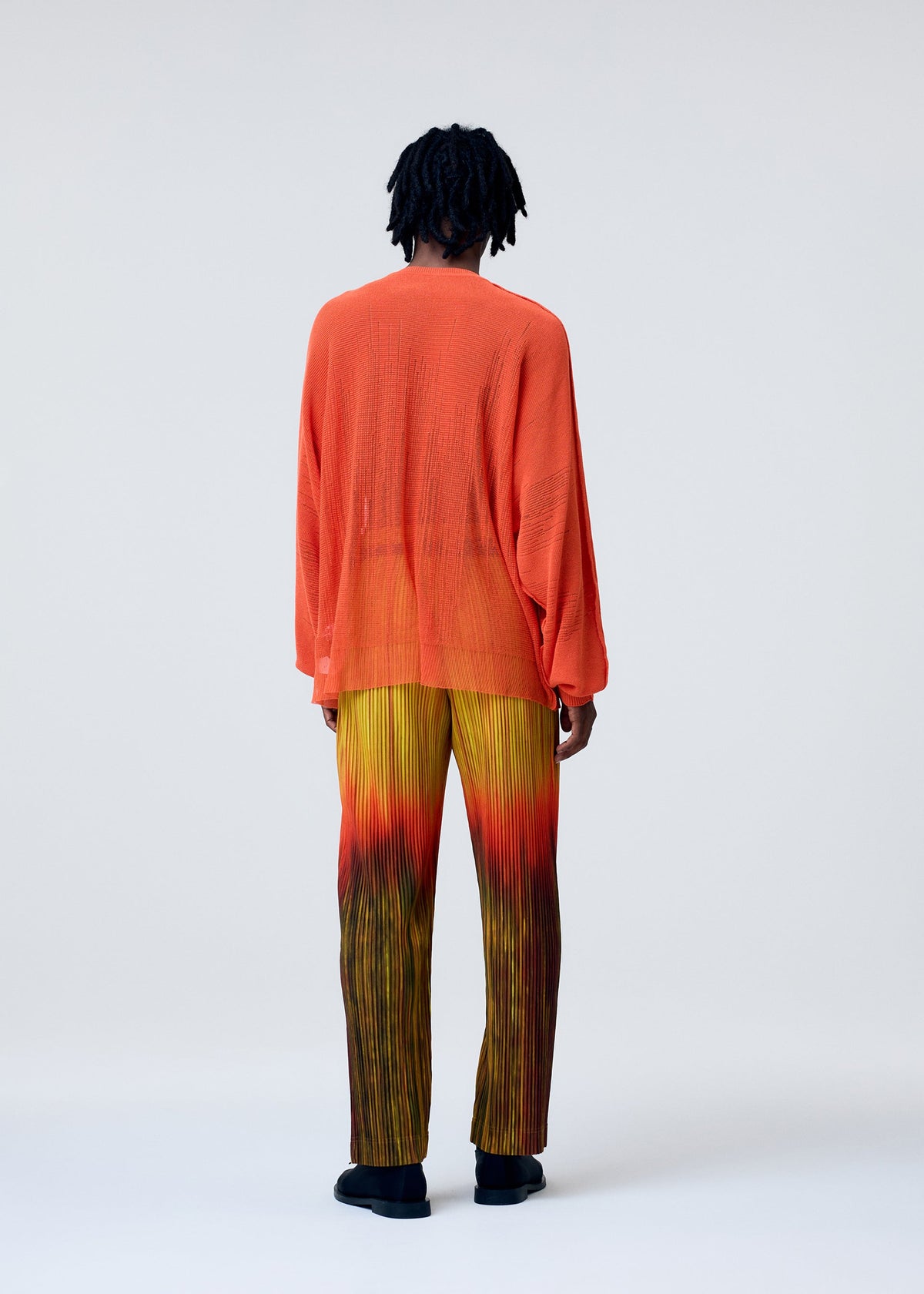 A person faces away, wearing the BRUSH CLOSE-UP KNIT by HOMME PLISSÉ ISSEY MIYAKE—a sheer orange long-sleeved top paired with pleated yellow-to-red gradient pants. The background is light and plain. view 6 of 7
