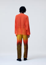 A person faces away, wearing the BRUSH CLOSE-UP KNIT by HOMME PLISSÉ ISSEY MIYAKE—a sheer orange long-sleeved top paired with pleated yellow-to-red gradient pants. The background is light and plain.