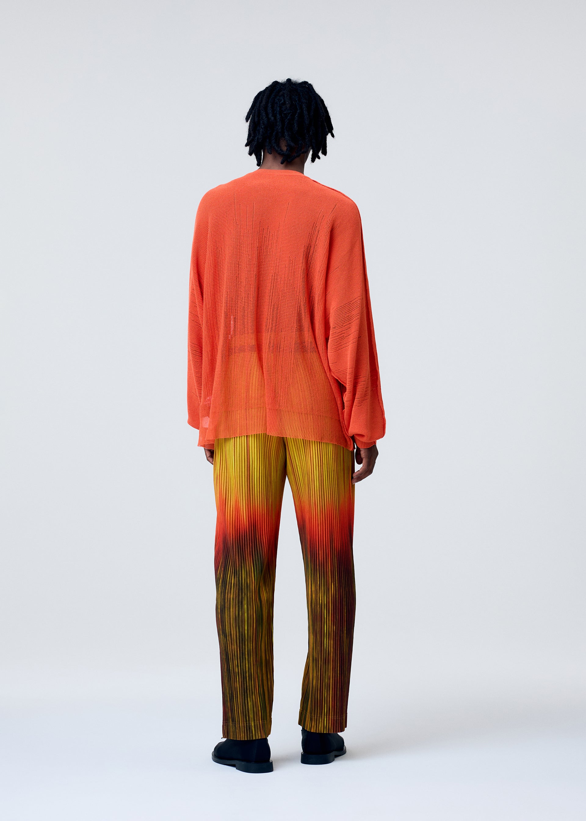Zoomed view A person faces away, wearing the BRUSH CLOSE-UP KNIT by HOMME PLISSÉ ISSEY MIYAKE—a sheer orange long-sleeved top paired with pleated yellow-to-red gradient pants. The background is light and plain.