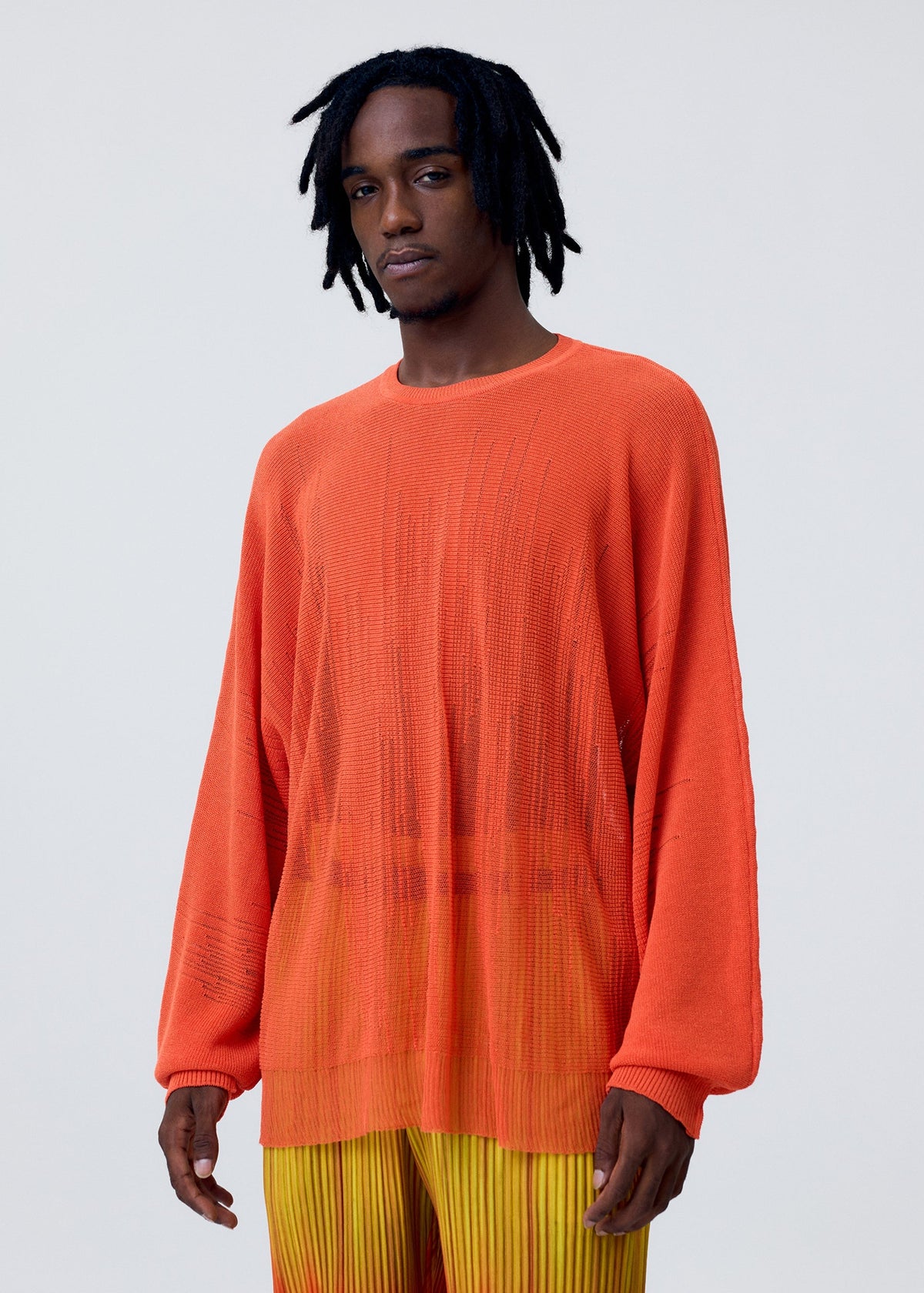 A man with short dreadlocks wears the BRUSH CLOSE-UP KNIT by HOMME PLISSÉ ISSEY MIYAKE—a long-sleeved, semi-sheer orange top—paired with yellow-orange pleated pants, standing against a plain light gray background. view 7 of 7