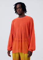 A man with short dreadlocks wears the BRUSH CLOSE-UP KNIT by HOMME PLISSÉ ISSEY MIYAKE—a long-sleeved, semi-sheer orange top—paired with yellow-orange pleated pants, standing against a plain light gray background.