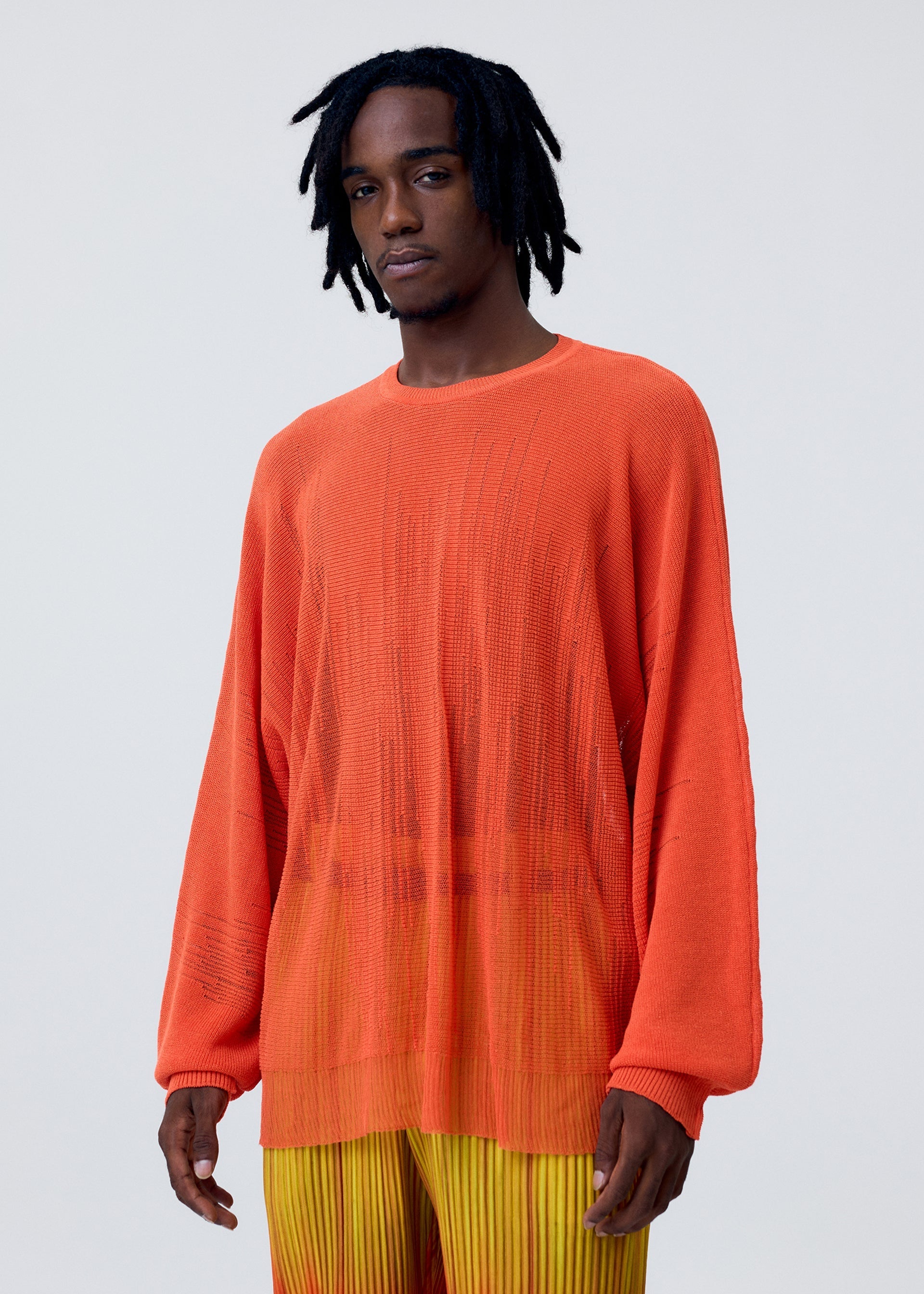 Zoomed view A man with short dreadlocks wears the BRUSH CLOSE-UP KNIT by HOMME PLISSÉ ISSEY MIYAKE—a long-sleeved, semi-sheer orange top—paired with yellow-orange pleated pants, standing against a plain light gray background.