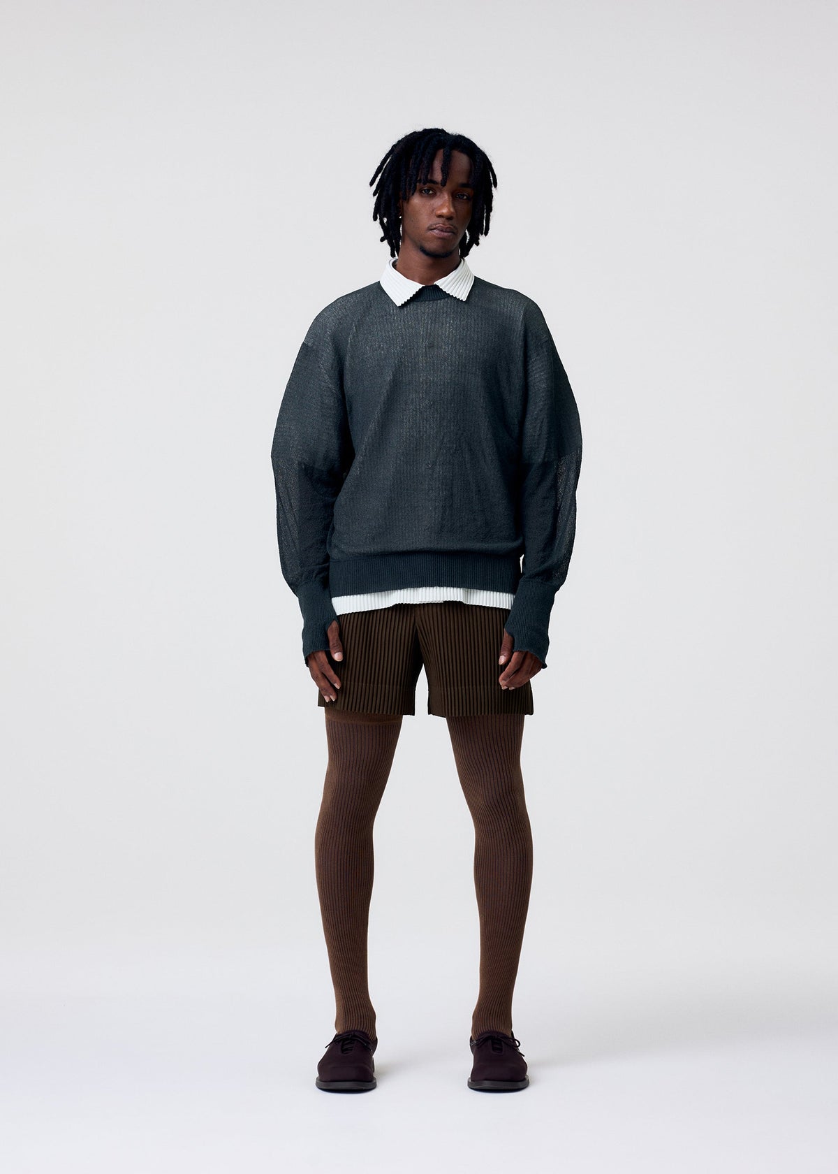 A person with short dreadlocks wears the HOMME PLISSÉ ISSEY MIYAKE KNIT UNRAVELED sweater over a white collared shirt, paired with brown shorts, ribbed tights, and dark shoes against a plain light background. view 6 of 8