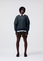 A person with short dreadlocks wears the HOMME PLISSÉ ISSEY MIYAKE KNIT UNRAVELED sweater over a white collared shirt, paired with brown shorts, ribbed tights, and dark shoes against a plain light background.