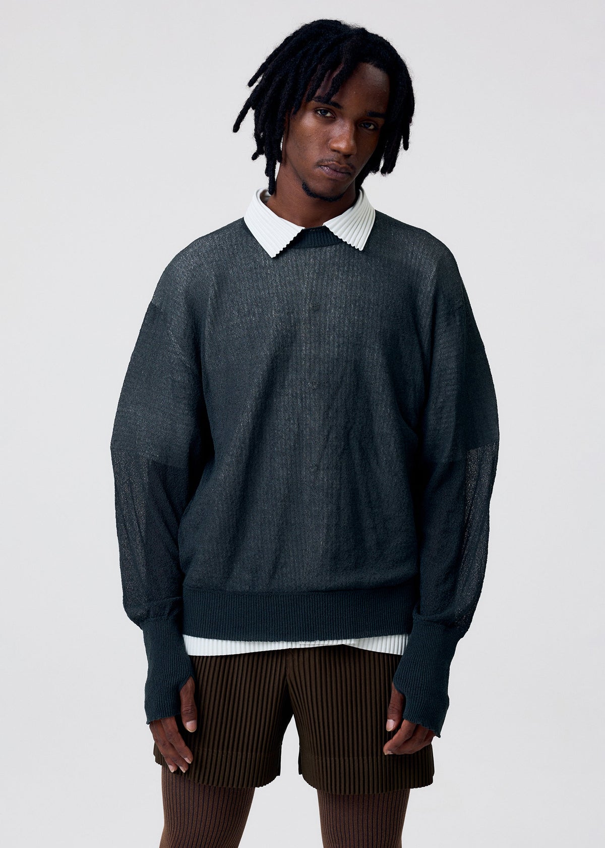 Wearing the KNIT UNRAVELED top by HOMME PLISSÉ ISSEY MIYAKE over a white collared shirt with brown striped shorts, a person with short dreadlocks stands against a plain light background. view 8 of 8