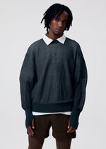 Wearing the KNIT UNRAVELED top by HOMME PLISSÉ ISSEY MIYAKE over a white collared shirt with brown striped shorts, a person with short dreadlocks stands against a plain light background.