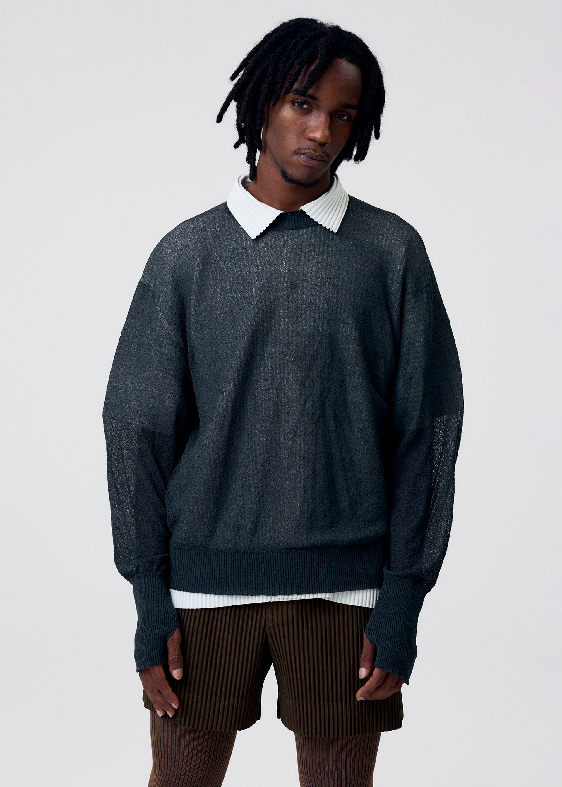 Zoomed view Wearing the KNIT UNRAVELED top by HOMME PLISSÉ ISSEY MIYAKE over a white collared shirt with brown striped shorts, a person with short dreadlocks stands against a plain light background.