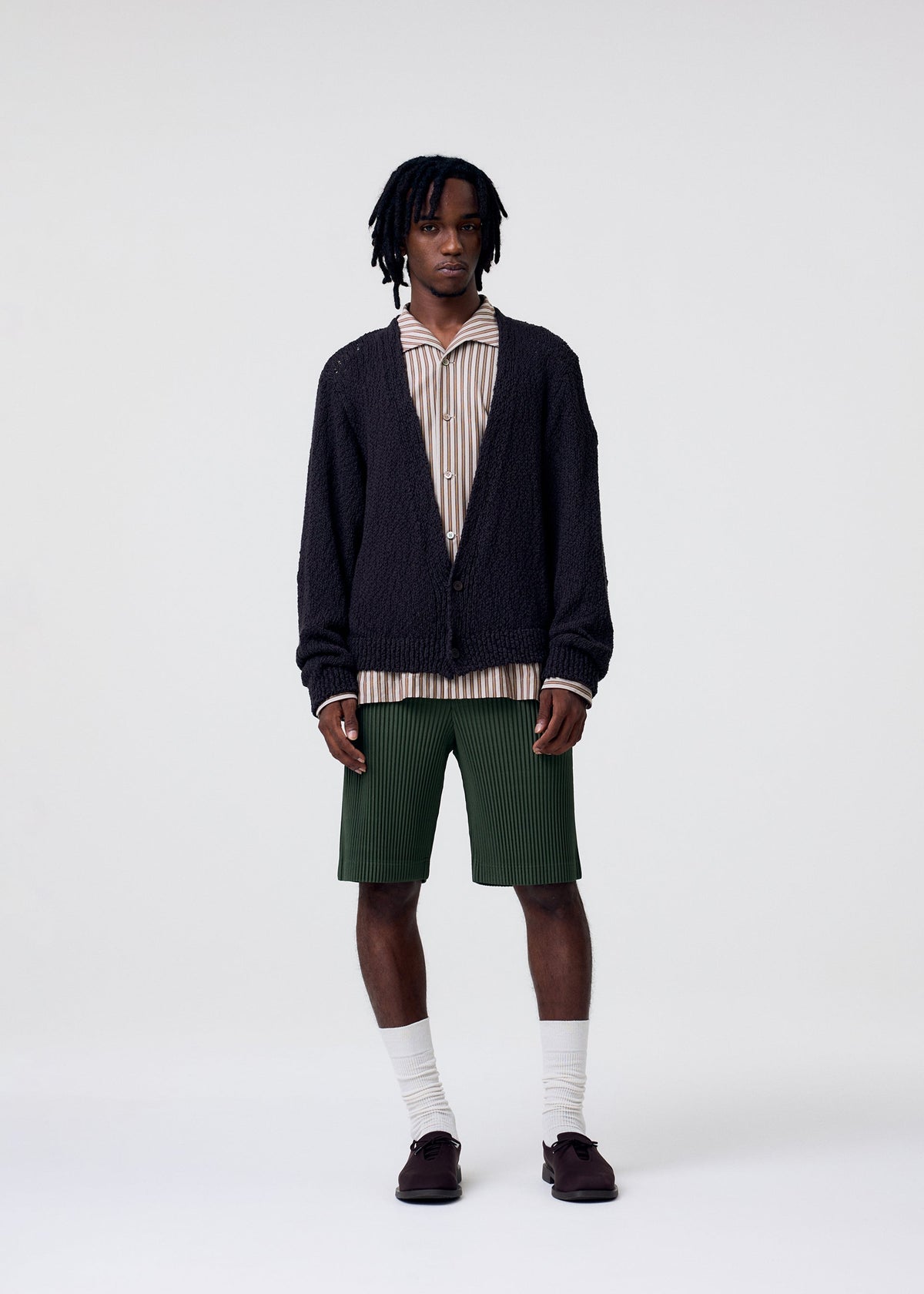 A man with short dreadlocks stands against a white background, wearing the RISOTTO KNIT by HOMME PLISSÉ ISSEY MIYAKE over a striped shirt with green shorts, white socks, and dark shoes for an effortless, casual style. view 6 of 8