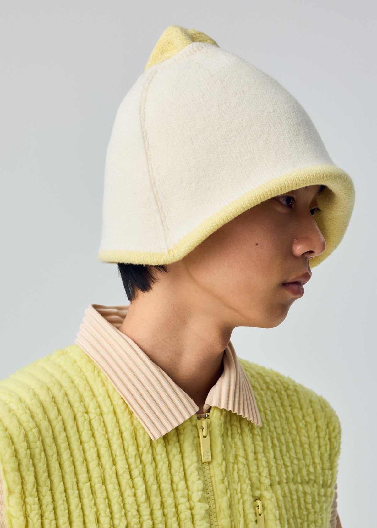 Zoomed view A person in WINTER GEAR : COLOR BLOCK by HOMME PLISSÉ ISSEY MIYAKE—a textured yellow jacket and a cream reversible beanie with yellow trim—seen in profile against a plain background.