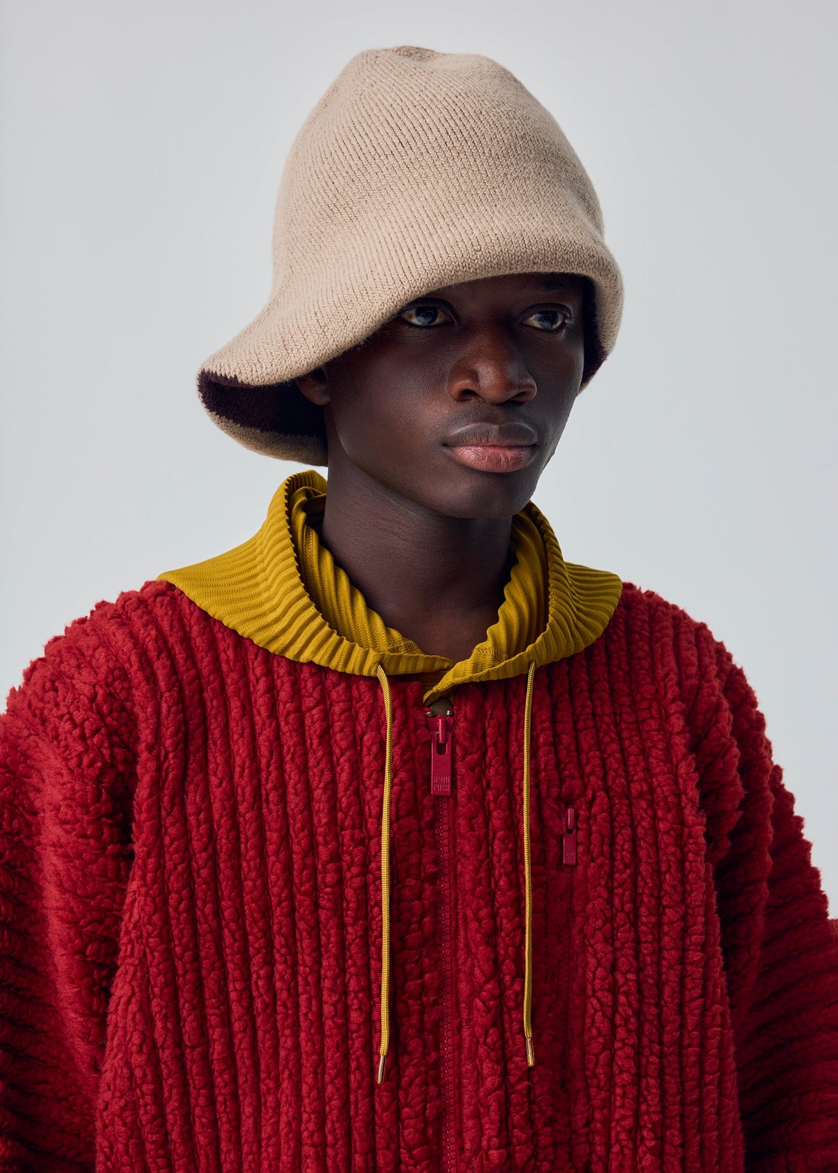 Zoomed view Wearing the WINTER GEAR : COLOR BLOCK by HOMME PLISSÉ ISSEY MIYAKE—a beige knit hat and a cozy red sweater with a yellow collar—this person gazes slightly off to the side against a plain background.
