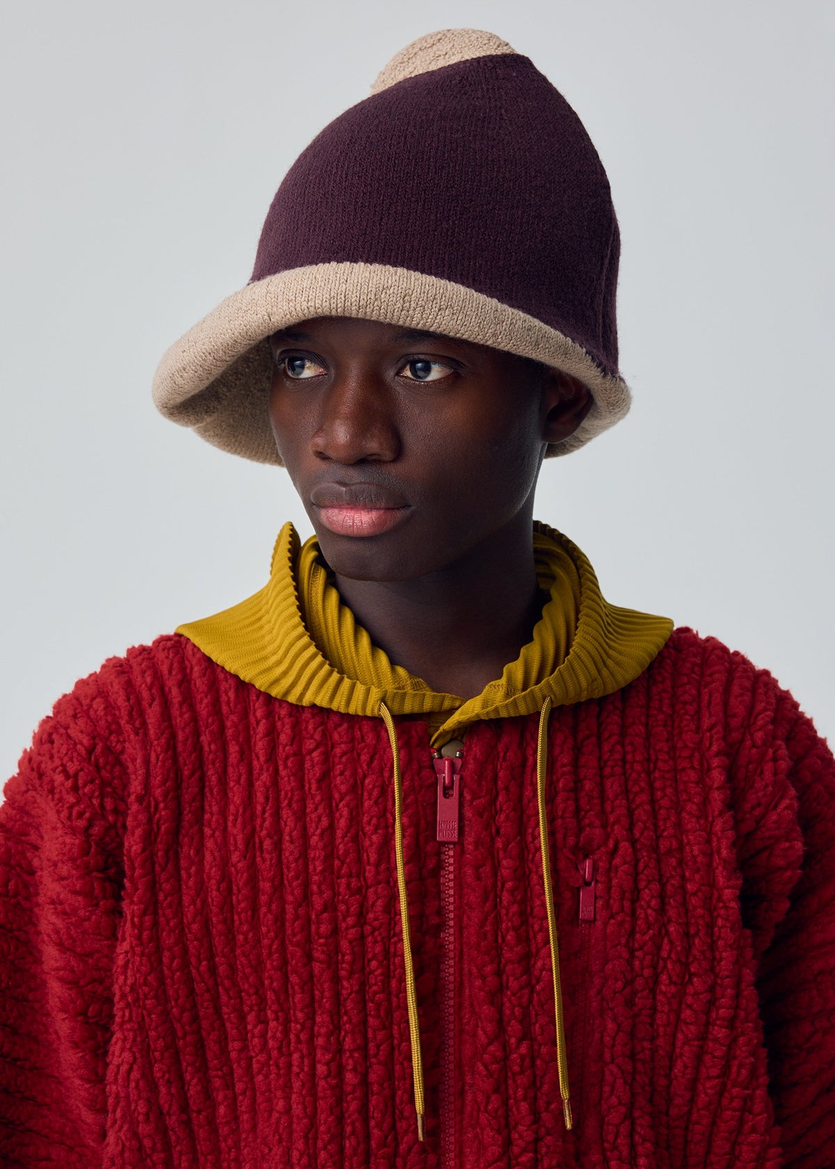 A person in the WINTER GEAR : COLOR BLOCK by HOMME PLISSÉ ISSEY MIYAKE—a textured red jacket with a yellow collar and a two-toned knitted hat—gazes to the side against a plain background. view 5 of 5