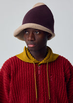 A person in the WINTER GEAR : COLOR BLOCK by HOMME PLISSÉ ISSEY MIYAKE—a textured red jacket with a yellow collar and a two-toned knitted hat—gazes to the side against a plain background.