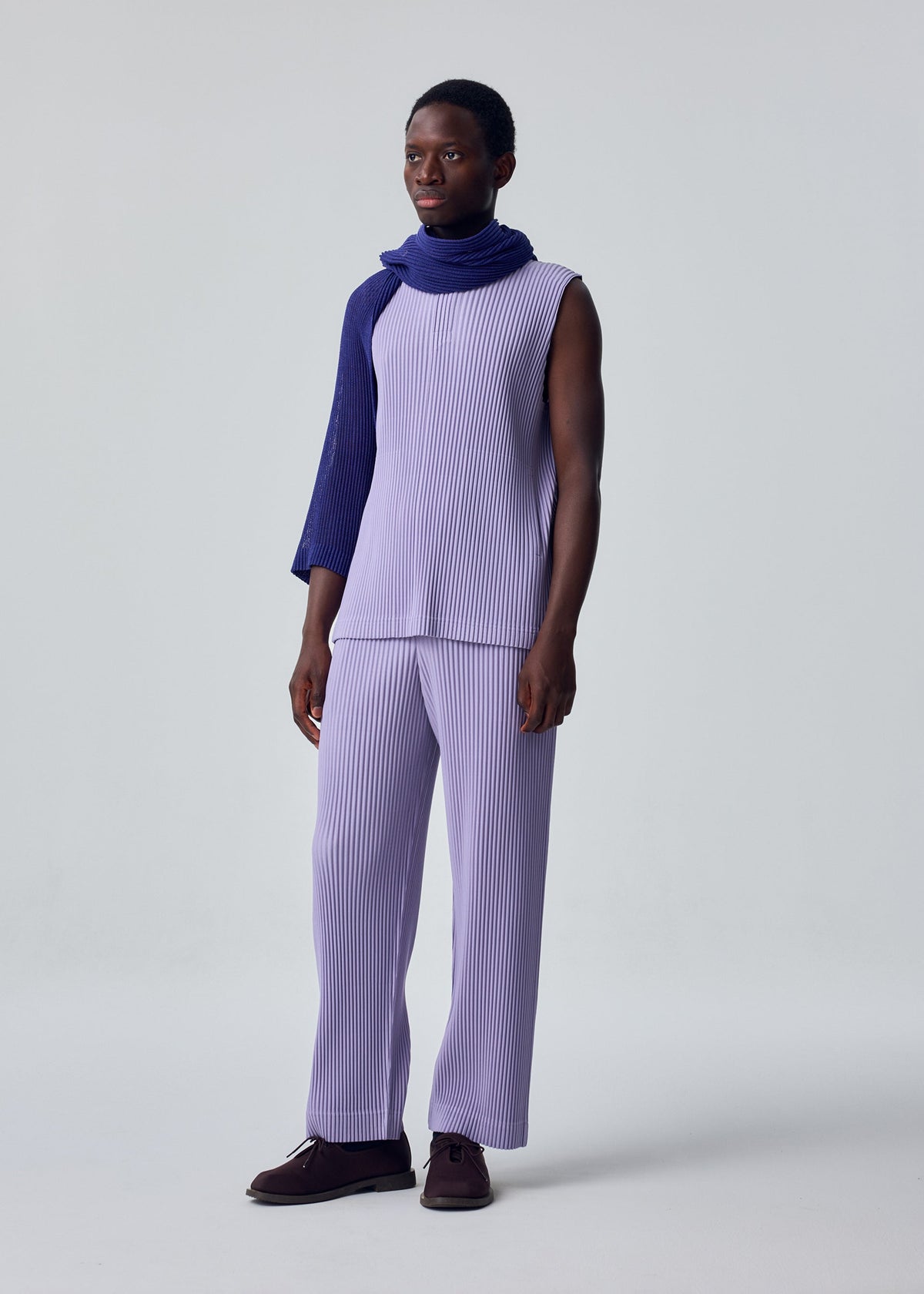 Against a plain backdrop, a person wears a sleeveless light purple pleated top with matching pants. A HOMME PLISSÉ ISSEY MIYAKE RUSTIC MESH STOLE in dark blue is draped around their neck. They wear dark shoes and smile slightly. view 4 of 6