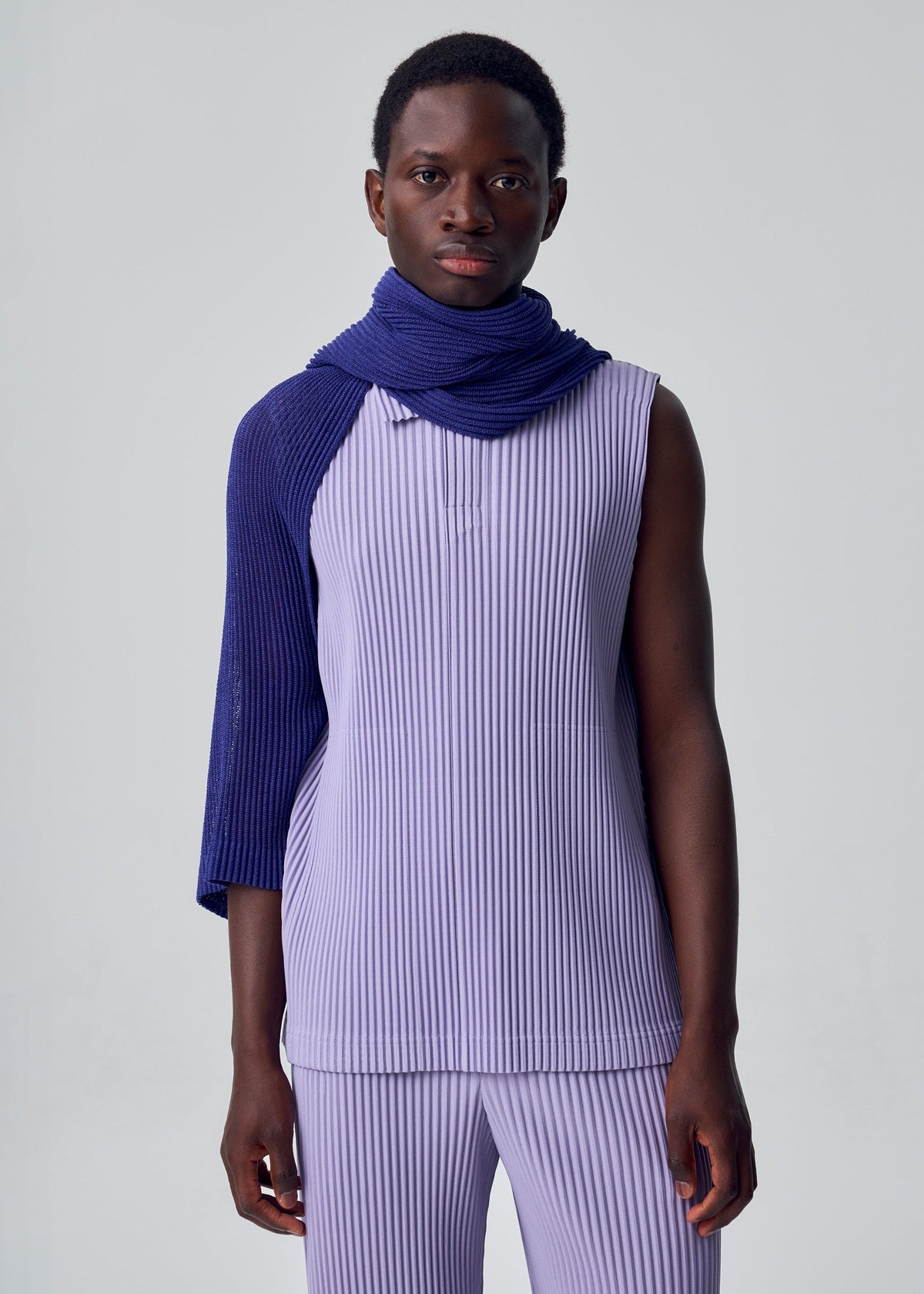 A person stands against a plain background, wearing a sleeveless, ribbed lavender top and matching pants, with the HOMME PLISSÉ ISSEY MIYAKE RUSTIC MESH STOLE in deep blue draped elegantly over one shoulder and around their neck. view 5 of 6