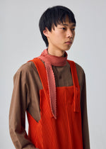 A person with short black hair poses against a plain, light gray background, wearing a brown shirt and a red pleated overall. As a striking neck accessory, they style the HOMME PLISSÉ ISSEY MIYAKE PLEATS STOLE—a red, striped scarf.