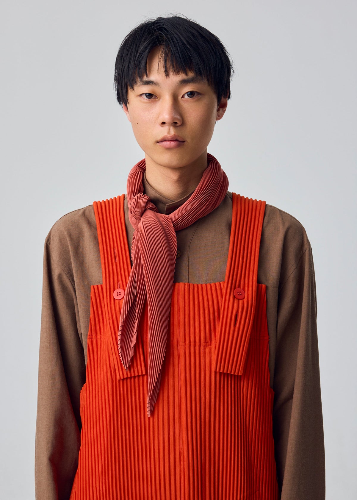 A person with short black hair stands against a plain light background, wearing a brown shirt and red pleated overalls with thick straps and buttons, styled with the HOMME PLISSÉ ISSEY MIYAKE PLEATS STOLE tied as a red scarf around the neck. view 6 of 6