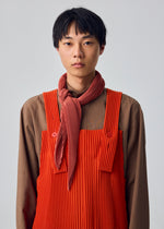 A person with short black hair stands against a plain light background, wearing a brown shirt and red pleated overalls with thick straps and buttons, styled with the HOMME PLISSÉ ISSEY MIYAKE PLEATS STOLE tied as a red scarf around the neck.