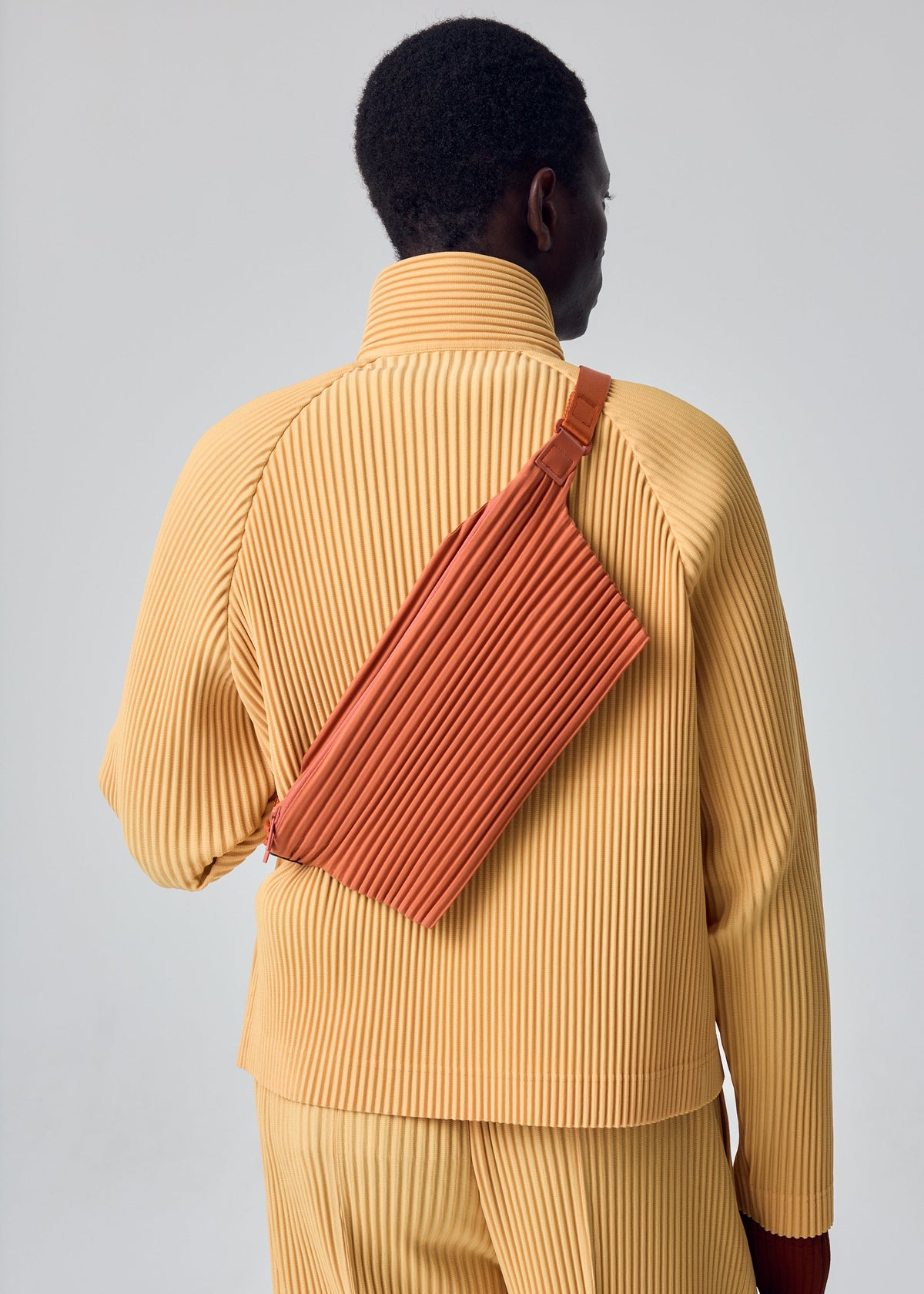 A person is shown from behind against a plain light background, wearing a pleated yellow outfit and carrying the HOMME PLISSÉ ISSEY MIYAKE PLEATS SLIM BAG in rust over their shoulder. view 5 of 6