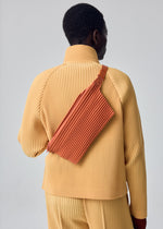 A person is shown from behind against a plain light background, wearing a pleated yellow outfit and carrying the HOMME PLISSÉ ISSEY MIYAKE PLEATS SLIM BAG in rust over their shoulder.