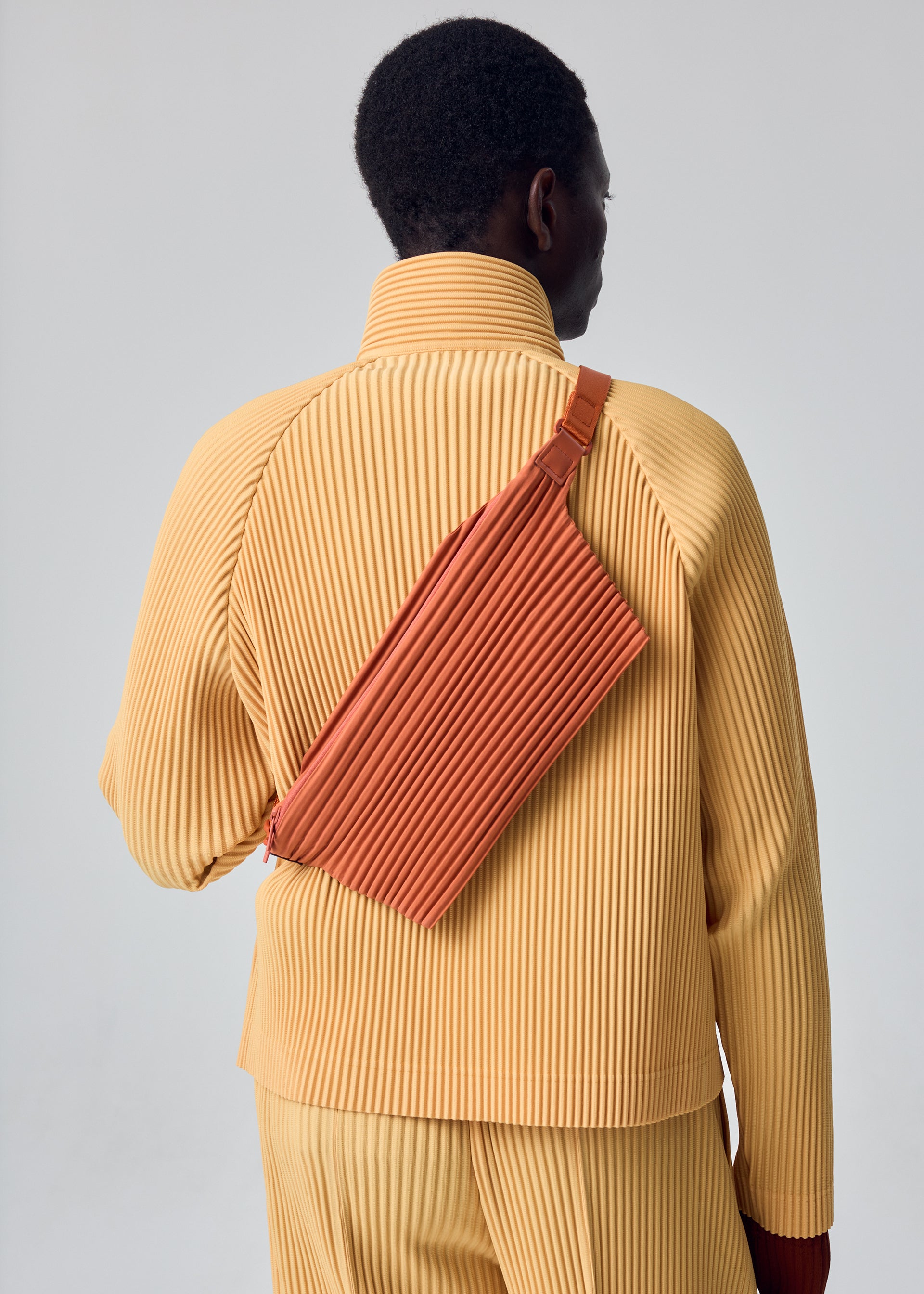 Zoomed view A person is shown from behind against a plain light background, wearing a pleated yellow outfit and carrying the HOMME PLISSÉ ISSEY MIYAKE PLEATS SLIM BAG in rust over their shoulder.