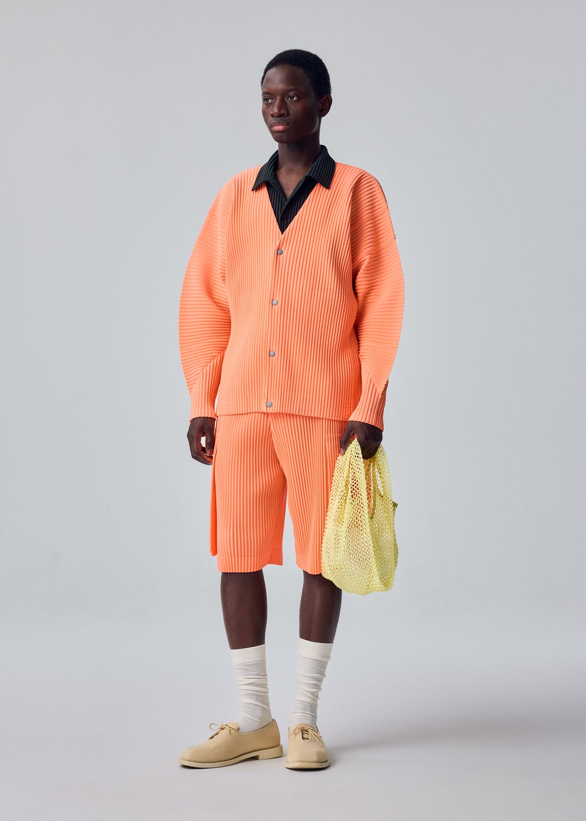 A person stands against a plain light background, showcasing a minimalist look featuring an orange pleated polyester cardigan and matching shorts, a black collared shirt, beige shoes with white socks, and carrying the MESH PLEATS BAG by HOMME PLISSÉ ISSEY MIYAKE. view 6 of 7