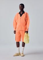 A person stands against a plain light background, showcasing a minimalist look featuring an orange pleated polyester cardigan and matching shorts, a black collared shirt, beige shoes with white socks, and carrying the MESH PLEATS BAG by HOMME PLISSÉ ISSEY MIYAKE.